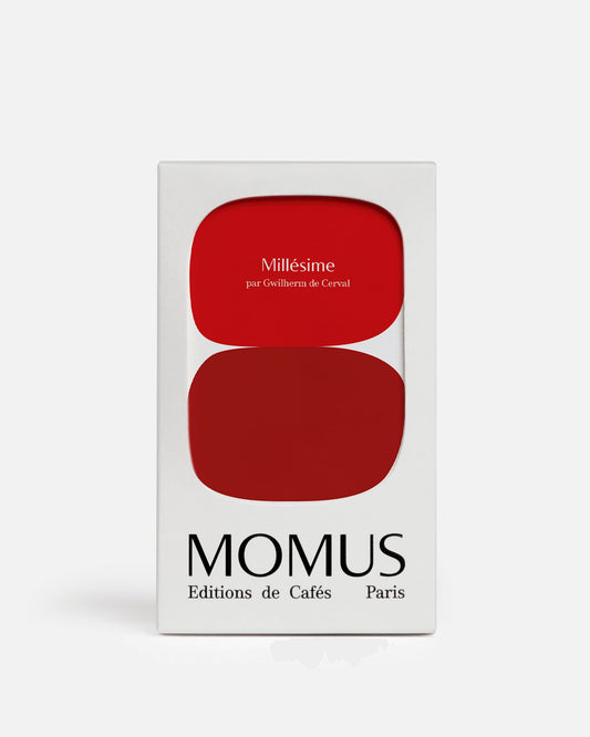 Gros plan du packaging blanc de l'édition spéciale Café Millésime de Momus. La boîte présente deux formes rouges superposées et arrondies. Le texte sur le rouge supérieur indique "Millésime par Gwilherm de Cerval". Le bas de la boîte porte la marque MOMUS Éditions de Cafés Paris. Café biodynamie vieilli.