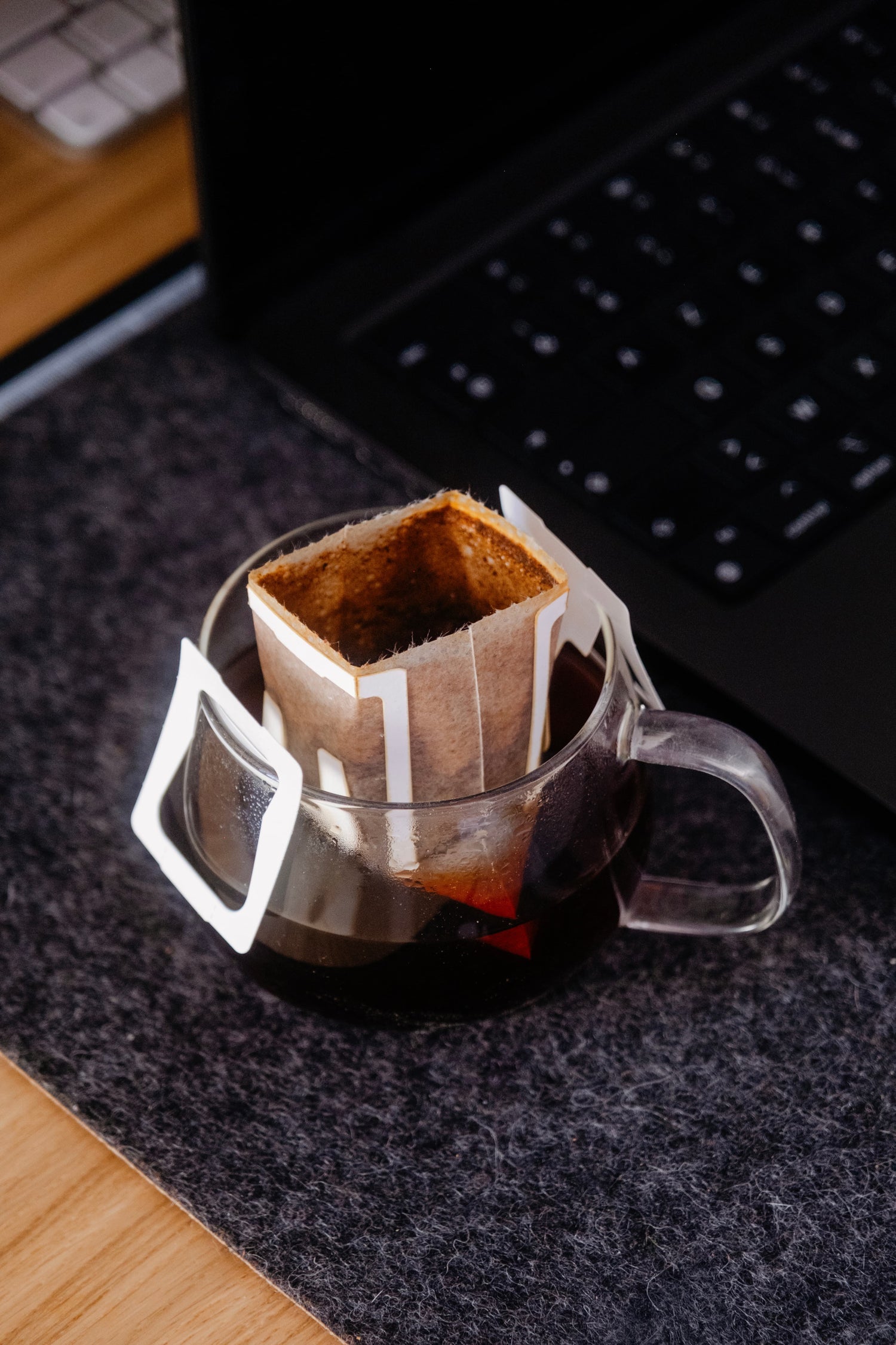 Sachet de café filtre individuel en infusion dans une tasse en verre sur un tapis de bureau, avec clavier d'ordinateur portable visible en arrière-plan