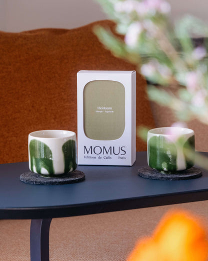 Packaging café Momus Paris gamme Heirloom Éthiopie présenté avec deux tasses artisanales à motifs végétaux verts sur table d'appoint moderne