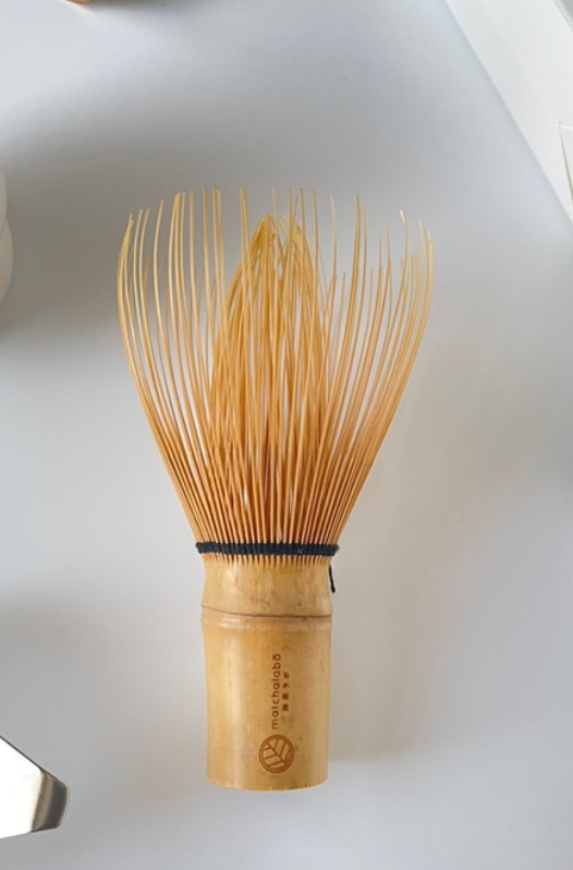 Fouet à matcha traditionnel japonais en bambou (chasen) avec fines tiges évasées, posé sur assiette blanche minimaliste