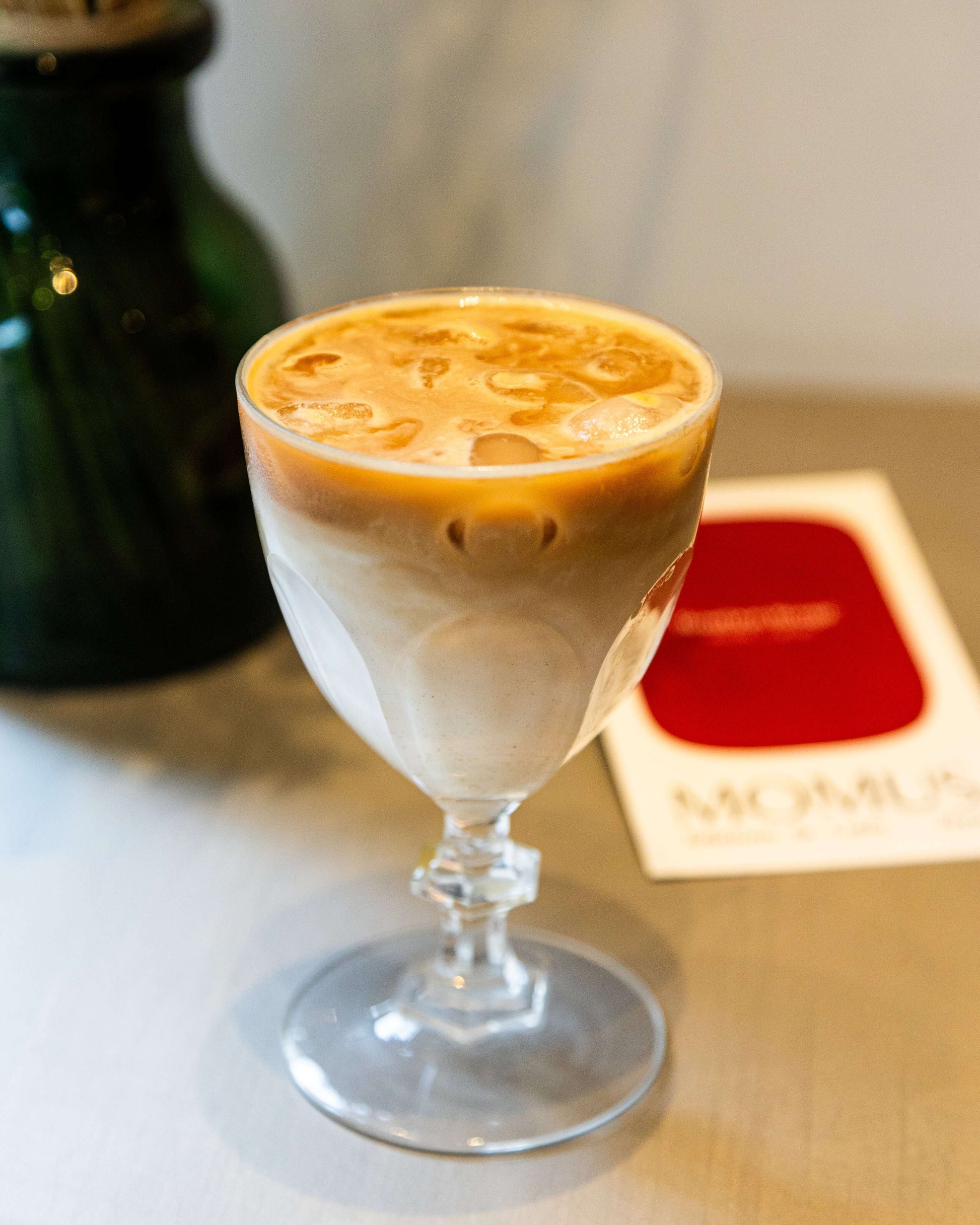 Gros plan sur un Iced Latte à deux tons, servi dans un verre à pied élégant. On aperçoit les couches de lait et d'espresso sur glaçons, avec l'emballage "Bourbon Rouge" de Momus en arrière-plan flou.
