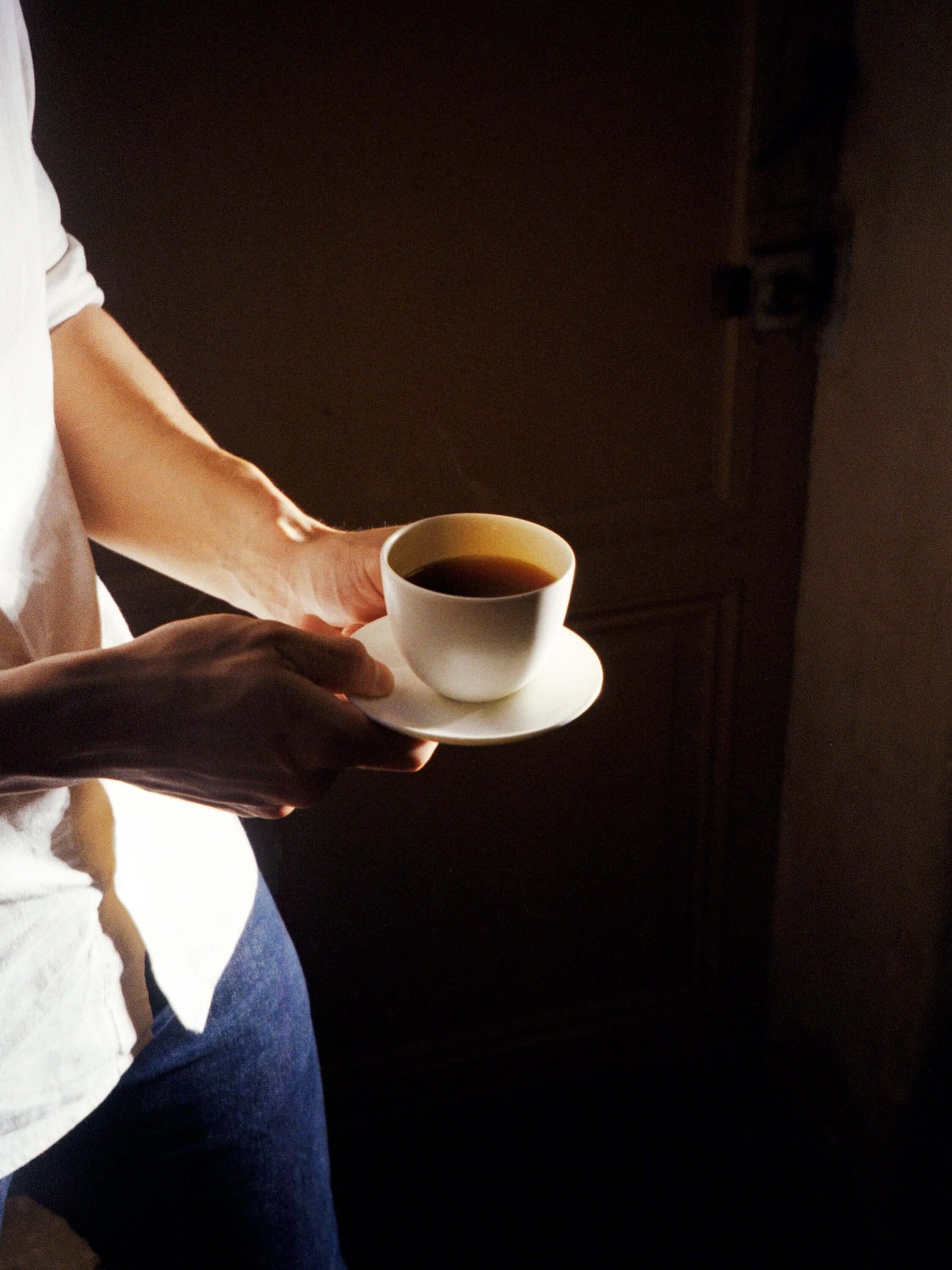 Deux mains tenant une tasse de café noir et sa soucoupe, éclairées par un rai de lumière sur le côté gauche. La personne porte une chemise blanche et un jean. Le fond est sombre.