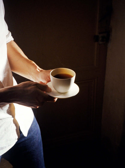 Deux mains tenant une tasse de café noir et sa soucoupe, éclairées par un rai de lumière sur le côté gauche. La personne porte une chemise blanche et un jean. Le fond est sombre.