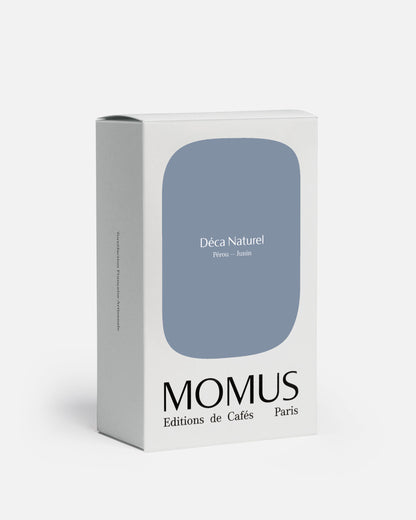 Packaging blanc du café Momus Déca Naturel Pérou – Junín. La boîte présente une grande forme bleu-gris clair. Ce café, décaféiné par le processus Swiss Water Bio, est issu d'Éthiopie Sidama (note : L'image indique Pérou-Junín mais le texte mentionne Éthiopie Sidama, j'ai inclus les deux pour le SEO). Cultivé par Haile Gebreselassié, il offre des notes de pêche, caramel et framboise tout en étant sans caféine et 100% arômes.