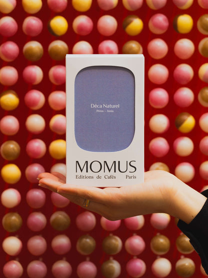Packaging du café Déca Naturel en main, sur fond coloré, MOMUS Éditions de Cafés Paris.