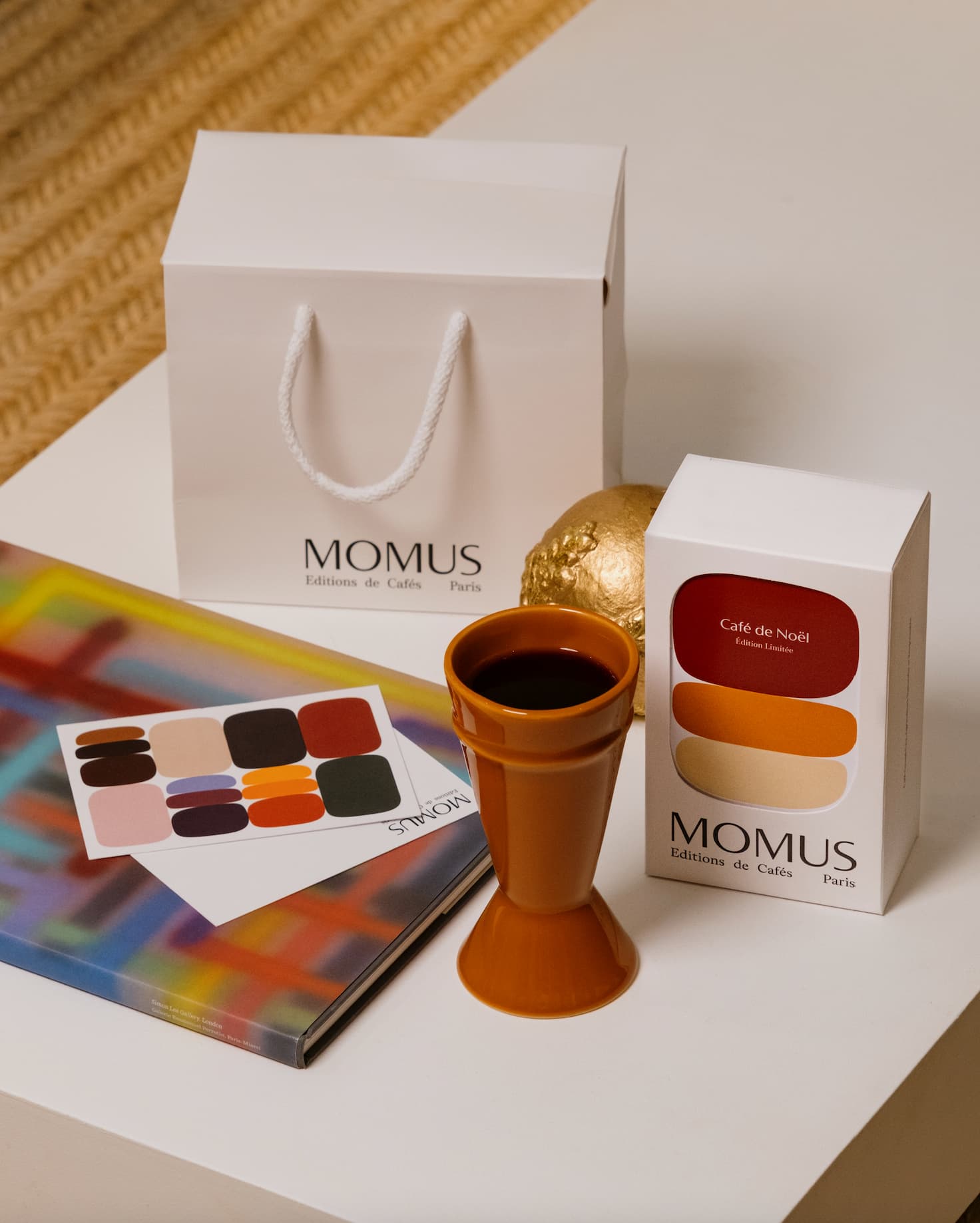 Coffret Café de Noël Momus présenté avec une tasse orange, un sac cadeau blanc, un livre coloré et une décoration dorée sur une table.