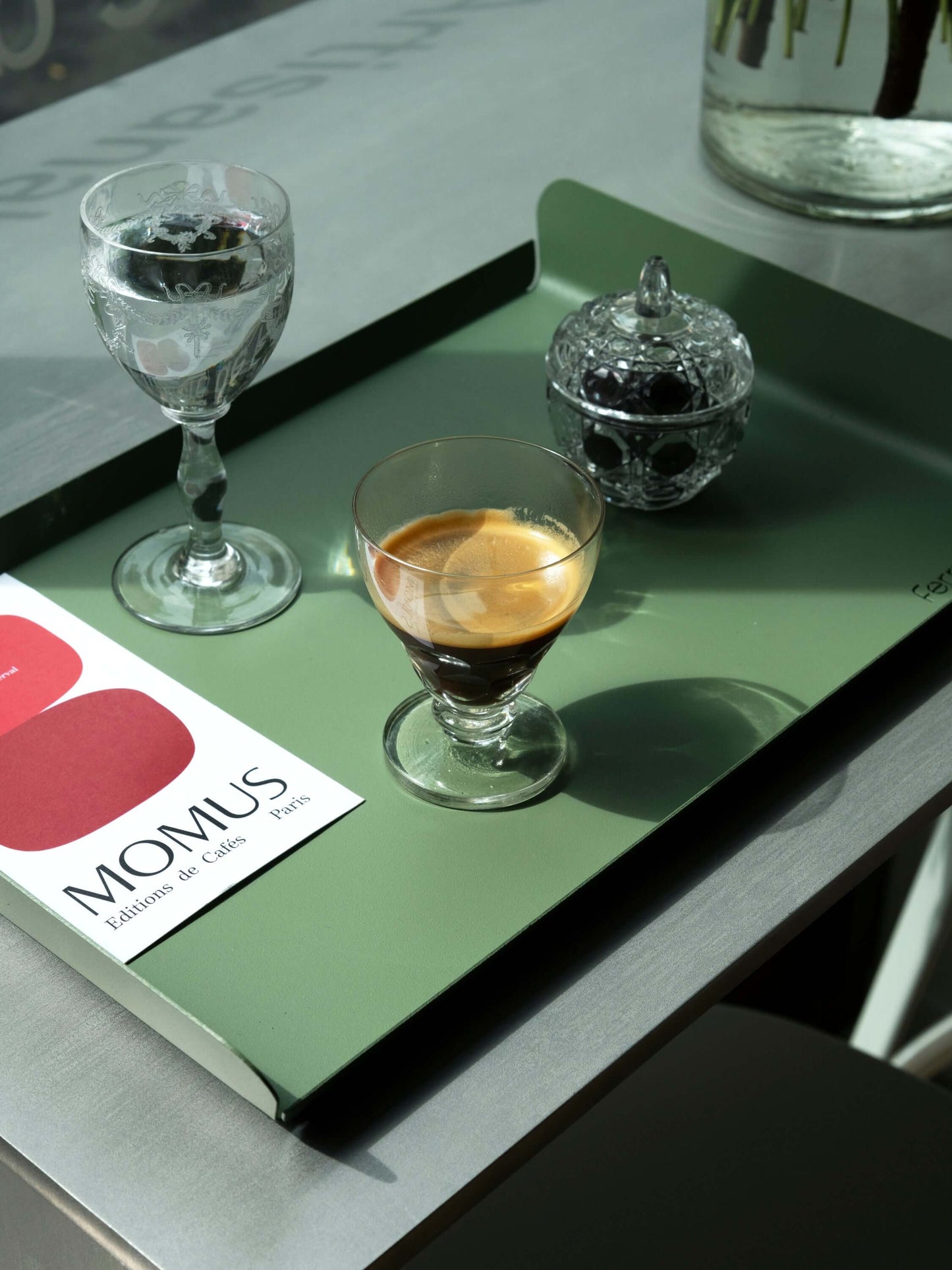 Un plateau vert olive posé sur une table métallique. Le plateau présente un verre d'eau, un verre d'espresso et une petite boîte à sucre en verre taillé. La carte de la omposition du café millésime est visible sur le plateau.