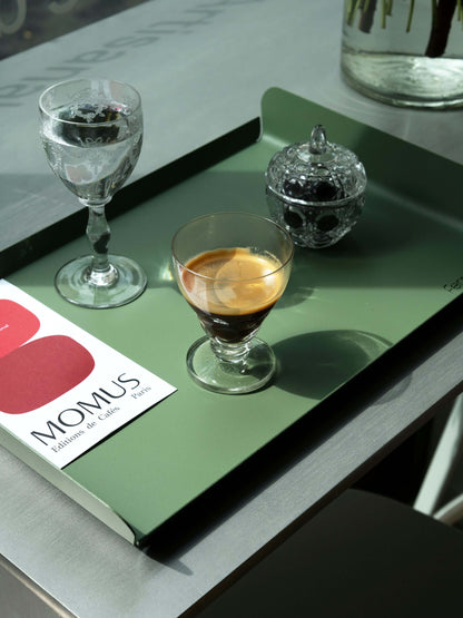 Un plateau vert olive posé sur une table métallique. Le plateau présente un verre d'eau, un verre d'espresso et une petite boîte à sucre en verre taillé. La carte de la omposition du café millésime est visible sur le plateau.