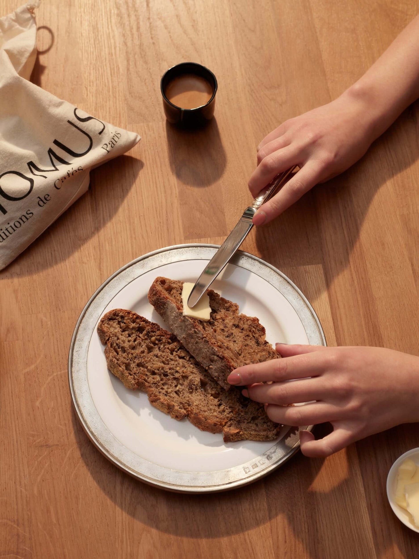 Vue plongeante sur deux mains tartinant du beurre sur des tranches de pain sur une assiette. Un petit sac Momus et une tasse de café sont posés sur la table en bois.