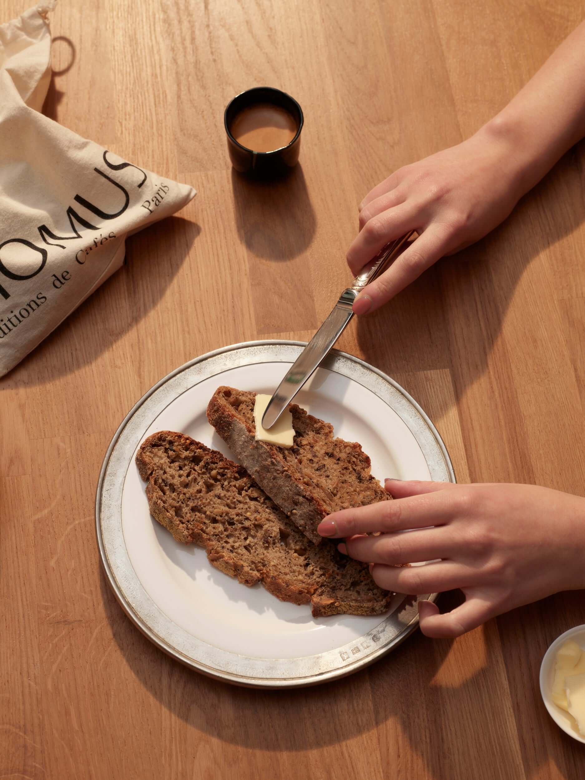 Vue plongeante sur deux mains tartinant du beurre sur des tranches de pain sur une assiette. Un petit sac Momus et une tasse de café sont posés sur la table en bois.
