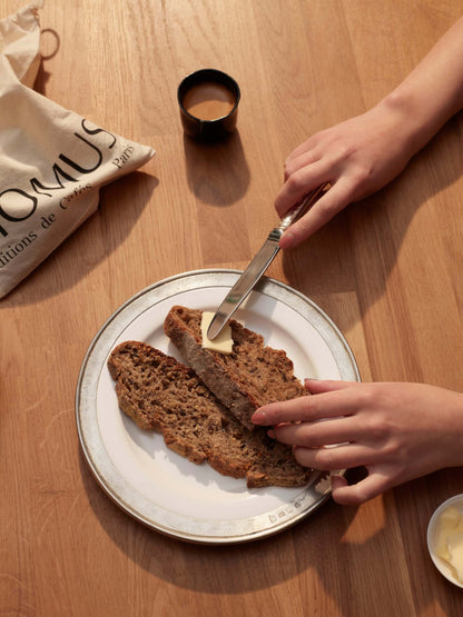Vue plongeante sur deux mains tartinant du beurre sur des tranches de pain sur une assiette. Un petit sac Momus et une tasse de café sont posés sur la table en bois.