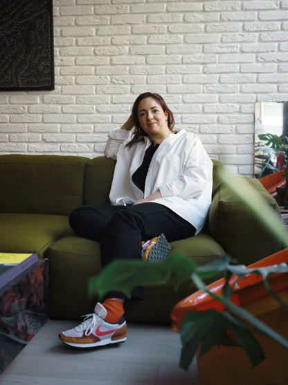 Portrait en plan large de Daniela Capuano, MOF, assise décontractée sur un canapé vert olive. Elle est vêtue d'une chemise blanche ample ouverte, d'un haut noir, d'un pantalon noir, de chaussettes orange vif et de baskets de style rétro blanc, rouge et marron. 