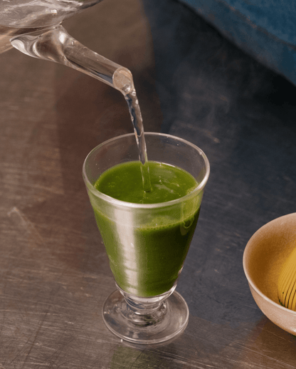 Versement d’eau chaude dans un verre contenant du thé matcha fraîchement fouetté, pour une boisson verte onctueuse et énergisante.