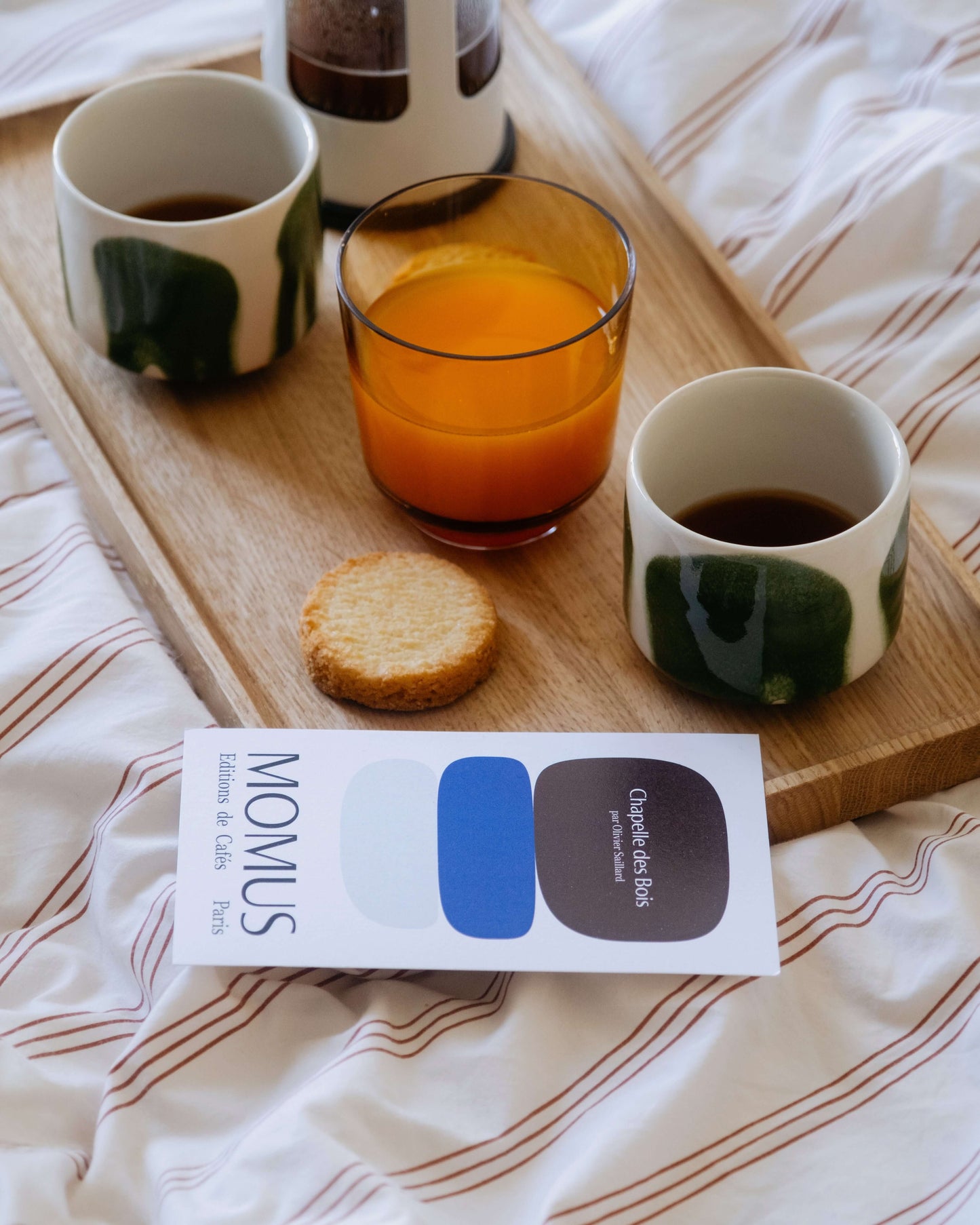Mise en scène petit-déjeuner cosy avec cafés Momus servis dans des tasses artisanales, accompagnés de jus d'orange frais et biscuit sur plateau en bois