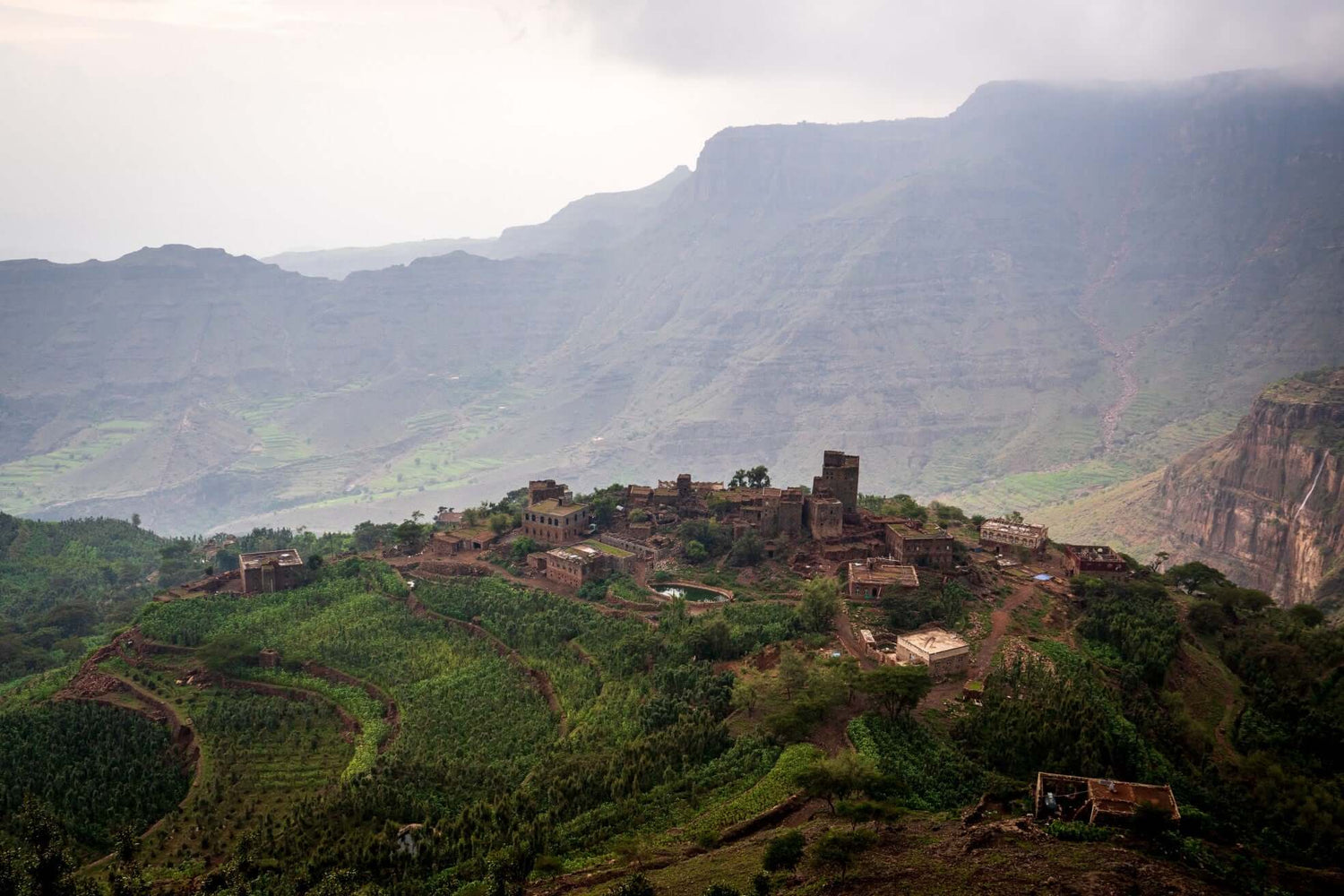 Village traditionnel,Yemenia, en pierre perché sur colline verdoyante entouré de cultures en terrasses, avec massif montagneux escarpé et vallée brumeuse en arrière-plan