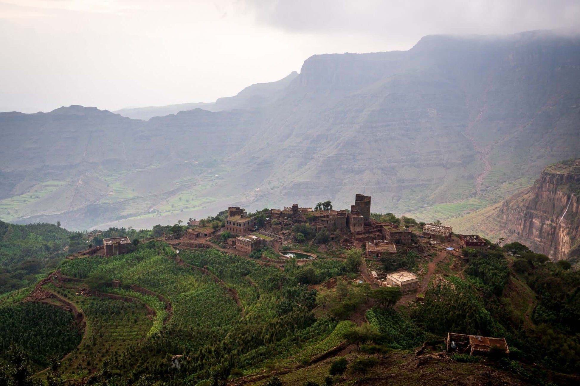 Village traditionnel,Yemenia, en pierre perché sur colline verdoyante entouré de cultures en terrasses, avec massif montagneux escarpé et vallée brumeuse en arrière-plan